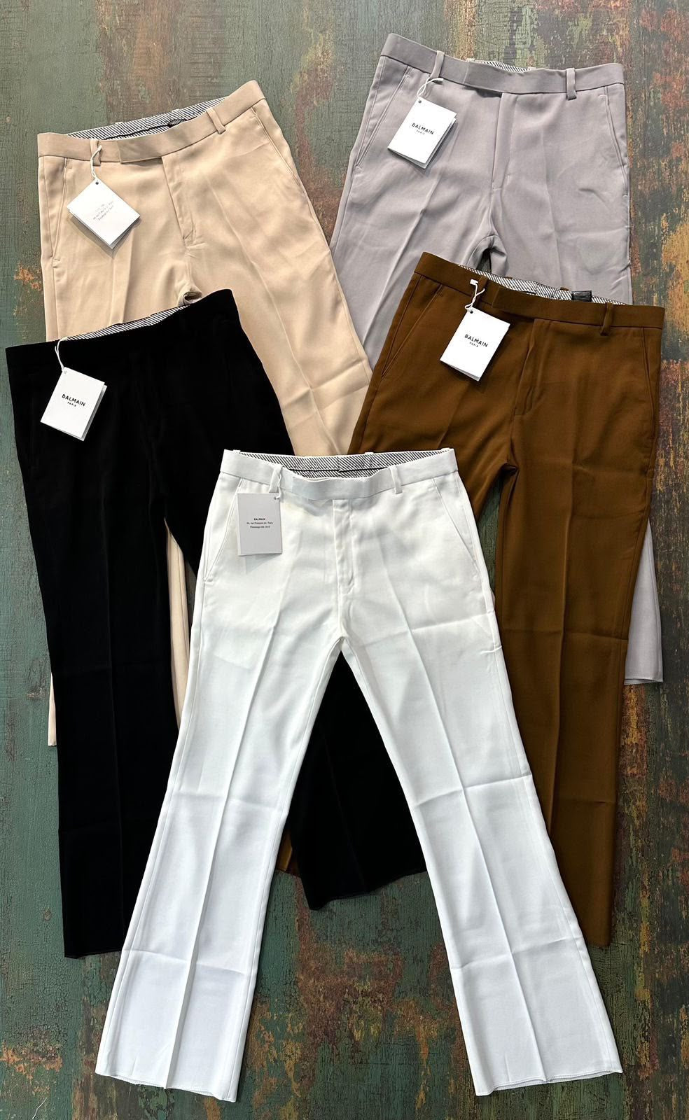 Six pairs of women's tailored trousers in different colors (white, black, beige, light gray, brown, and taupe) are neatly folded and arranged in a fan-like layout on a rustic, textured green and brown wooden surface. Each pair has a white paper tag attached to the waistband.